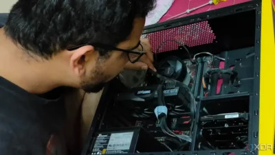 View of an open PC case showing cards and fan inside with cables connecting everything. A man has his face close to the case, and is using one finger to pull a black cable slightly away from a fan on the motherboard.