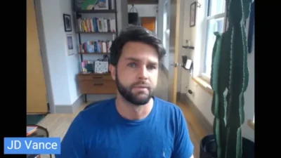 A webcam view of a man wearing a blue t-shirt, sitting in a room with an open door behind him and a bookcase with books. To the right stands a green cactus type plant. The man's face looks like JD Vance.