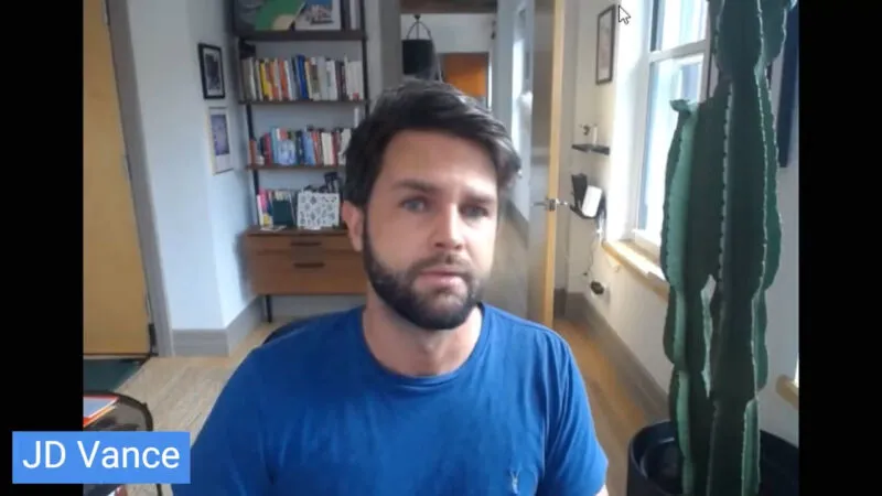 A webcam view of a man wearing a blue t-shirt, sitting in a room with an open door behind him and a bookcase with books. To the right stands a green cactus type plant. The man's face looks like JD Vance.