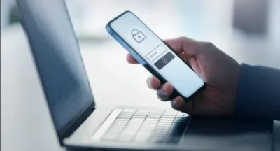 Side close-up view of an open laptop. Above it hovers a hand holding a smartphone, showing a login screen on it.