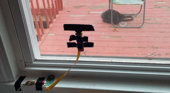 View through a window. On the window pane is taped a thin rectangular device with a brown data cable hanging down from it. In the background is a red wooden deck with a folding chair. Under the chair sits a black cat hovering near a food bowl, and looking up at the window.