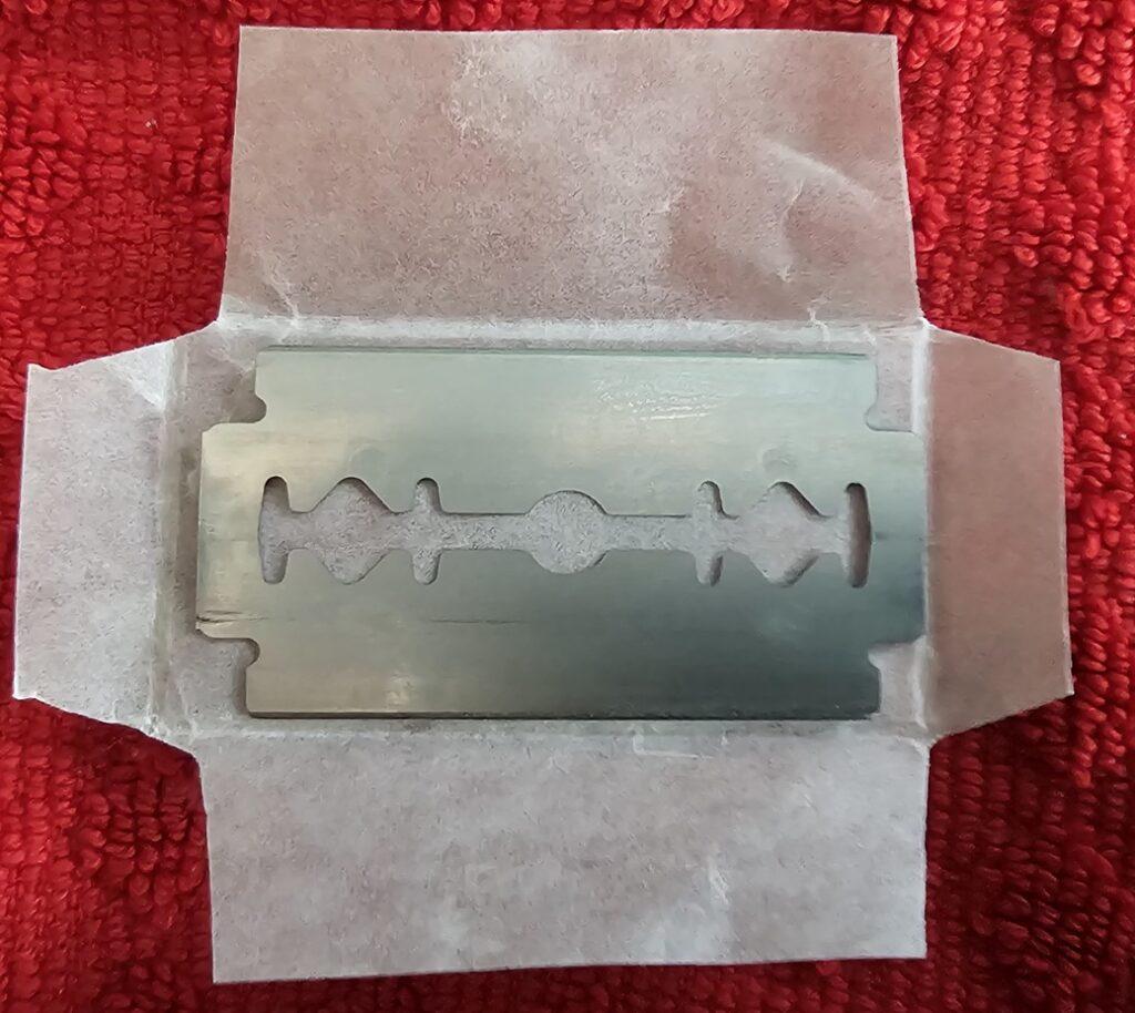 A silver coloured double edge razor blade with no printing on it at all. It is resting on its open paper packaging, which also has no printing on it.