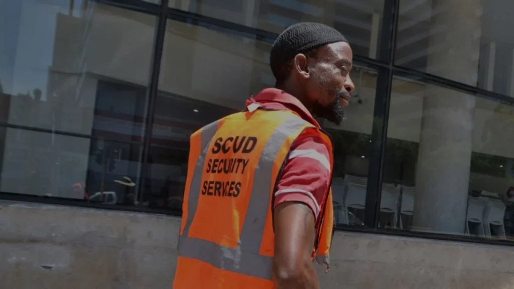 A car guard wearing a neon orange jacket with the words on the back: SCUD security services.