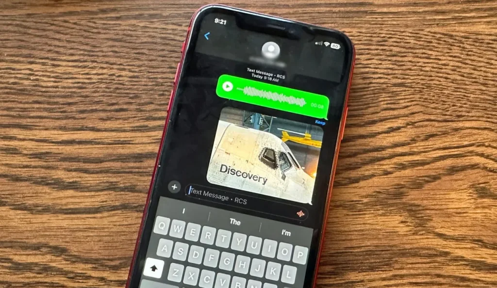 An iPhone resting on a wooden surface. On its screen can be seen the keyboard and an image of an aeroplane cockpit window with the word Discovery beneath it.