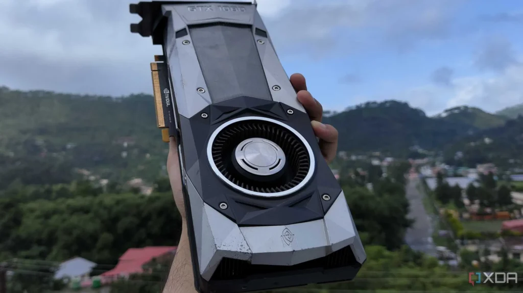 A silver coloured NVidia GTX 1080 graphics card being held in a hand. In the background is the blurred view of a town with some green trees and a hill in the background.