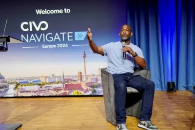 Man sitting in a chair on a stage, holding a microphone. Behind is a banner saying Welcome to Civo Navigate Europe 2024