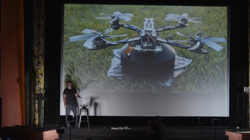 Man standing on a stage in front of a projector screen. ON the screen can be seen a drone resting on some grass. There are some extra wires protruding from the drone, and underneath it can be seen what looks like some flat discs protruding out around it.