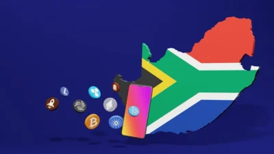 Outline of the borders of South Africa, with the centre filled in with the colours of the SA flag. To the left side is seen a blank outline of a smartphone, with various images of coins which have logos like Bitcoin and other crypto currencies.