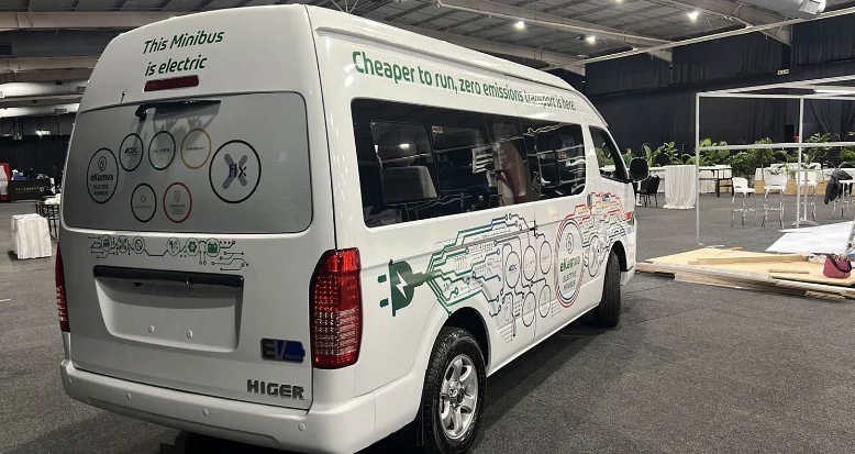 A white minibus taxi with words over the rear window stating "This Minibus is electric" and a model name seen is HIGER.