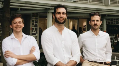 Three young men, all wearing white open neck shirts, standing outside of what looks like a restaurant in the background.