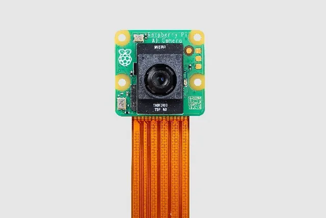 A Raspberry Pi camera module - square piece of green circuit board with a black camera mounted on it. Below is seen a brown ribbon cable.