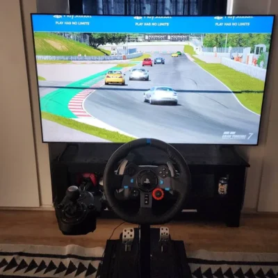 Large screen TV showing a car racing game with a few cars racing on a tar track. In front stands a black steering wheel, with a gear shifter on its left, and some foot pedals can be seen underneath it.