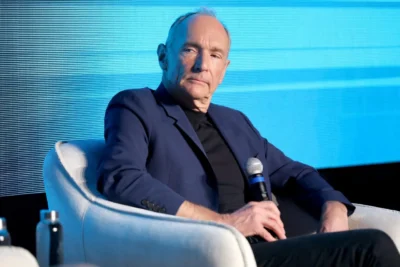 Man with thinning hair sitting ina chair on a podium holding a microphone in his right hand.