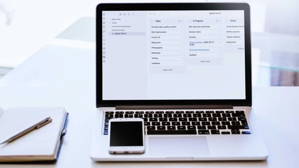 An open laptop showing a kanban layout project plan on the screen. On the keyboard rests a smartphone, and to the left of tye laptop is an open book with a pen lying on the page.