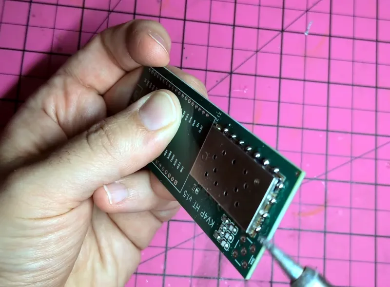 A hand holding a green coloured rectangle shaped circuit board, with a soldering iron tip poised over it.