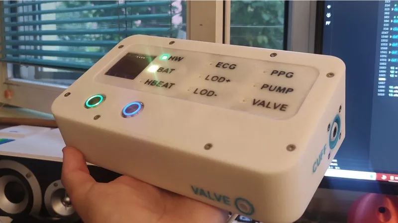 Rectangular white plastic box resting on the palm of a hand. It has slots on top for LED lights, and they are labelled HW, BAT, HBEAT, ECG, LOD+, LOD-, PPG, PUMP and VALVE. There is a green LED lit by HW. There are also green and blue coloured buttons below the LEDs.