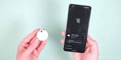 A hand holding a white disc shaped object. The other hand is holding a smartphone which has text on the screen saying Chipolo ONE Point and options for find nearby, play sound, and share device.