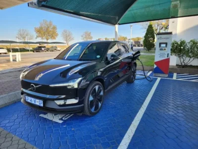 A black colour Volvo SUV parked next to an EV charging station. There is a black cable connected between the charging station and the rear left side of the car.