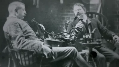 Two men in formal attire sitting in chairs next to a table. The men are each holding a tube with a funnel at the end towards their mouths. The tubes are connected to a mechanical device on the table. The device can be seen to have a large round cylinder on top, and a number of other metal rods and knobs.