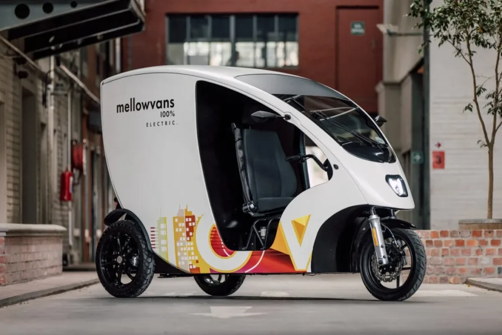 A three wheeled delivery van that is completely covered with an enclosed cargo area at the back. A sign on the side says mellowvans 100% electric.