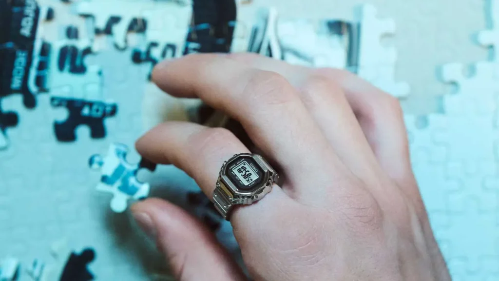 A hand showing a silver ring on the index finger. The ring is identical to a miniature Casio digital watch.