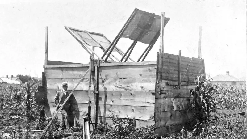 Old black and white hotos of a square wooden structure. The op is open, and two paddles of a wind turbine can be seen protruding. A man is standing in the front holding a wooden plank. He is wearing a boater hat and wearing a suite.