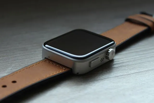 A square smartwatch. It has a metallic silver body with a black glass screen. On the side are a rotating know and a press button. Brown leather straps can be seen on both sides.