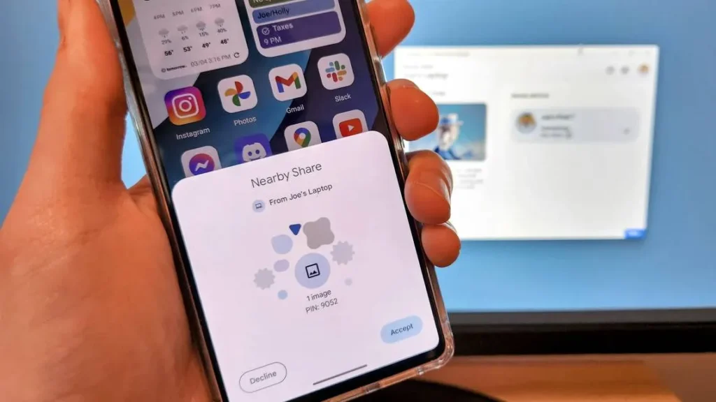 The image shows a hand holding a smartphone displaying a Nearby Share request from a laptop named "Joe's Laptop." The phone's screen shows various apps including Instagram, Photos, Gmail, and Slack. In the background, a computer monitor displays a blurred webpage or application. The foreground is dominated by the smartphone and hand, while the background is a simple, out-of-focus workspace. The location appears to be an office or home office setting. The PIN displayed on the phone, 9052, is unique to this specific file transfer request.