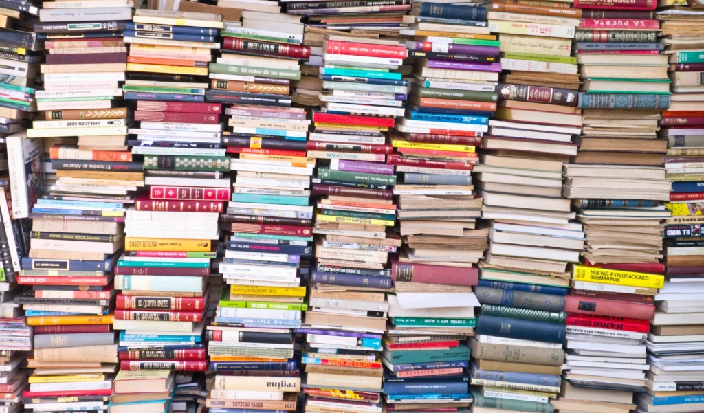 The image shows a wall of books stacked haphazardly, creating a visually stunning texture of various colors and sizes. The foreground is dominated by the densely packed books, while the background is mostly obscured by the sheer volume of literature. The location appears to be an outdoor book market, perhaps in a Spanish-speaking country, judging by some of the visible titles. The titles visible in the image show a mix of classic and contemporary literature, suggesting a wide diversity of literary preferences.