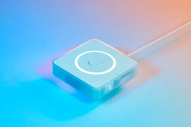 The image shows a small, square device, possibly a smart home gadget, resting on a surface. The background is a gradient of soft blue and orange hues, creating a calm and modern atmosphere. In the foreground, the device is the primary focus, with its sleek, white design and subtle glowing circle on the top drawing the viewer's attention. A thin cable is connected to the device, suggesting it is powered or connected to a network.