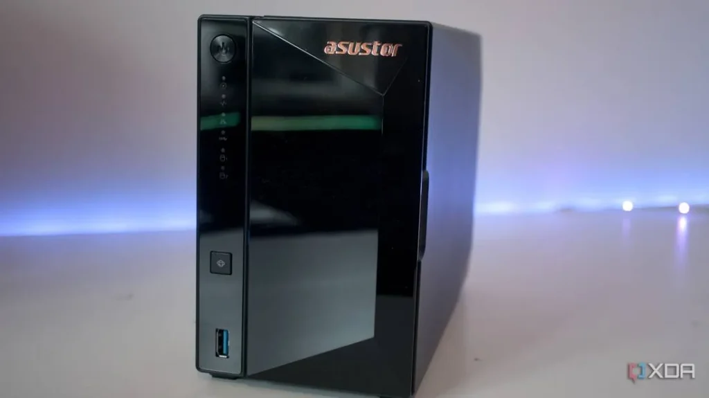 The image shows a black ASUSTOR NAS device in the foreground, which appears to be a Network Attached Storage device. The background features a blurred-out white surface, and there's a subtle blue light source emanating from the bottom, possibly from LED strip lighting. The overall setting looks like a product photography studio setup, indicating a professional product shot.