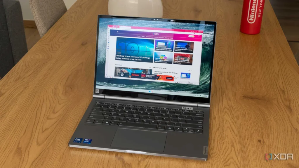 A Lenovo laptop is open on a light brown wooden table, showcasing a webpage about Windows 10's 10th anniversary. The background subtly reveals a portion of a living room, including what seems to be a grey couch and a wooden floor. In the foreground, besides the laptop, there is also a red Nintendo-branded water bottle with a slightly worn-out label.