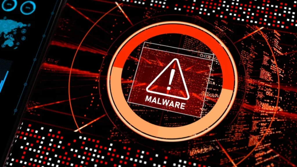 The image shows a close-up view of a digital screen displaying a malware alert. In the foreground, a prominent warning sign with an exclamation mark inside a triangle and the word "MALWARE" is clearly visible. This is encircled by a segmented, glowing orange ring, possibly representing a progress bar or severity indicator. The background is filled with a complex array of digital data streams, red and black in color, giving the impression of a computer network or system under stress. A portion of another screen, showing charts and a world map, is visible in the upper left corner, suggesting a broader technological context.