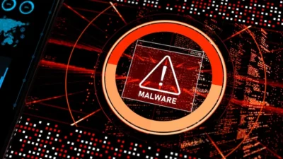 The image shows a close-up view of a digital screen displaying a malware alert. In the foreground, a prominent warning sign with an exclamation mark inside a triangle and the word "MALWARE" is clearly visible. This is encircled by a segmented, glowing orange ring, possibly representing a progress bar or severity indicator. The background is filled with a complex array of digital data streams, red and black in color, giving the impression of a computer network or system under stress. A portion of another screen, showing charts and a world map, is visible in the upper left corner, suggesting a broader technological context.