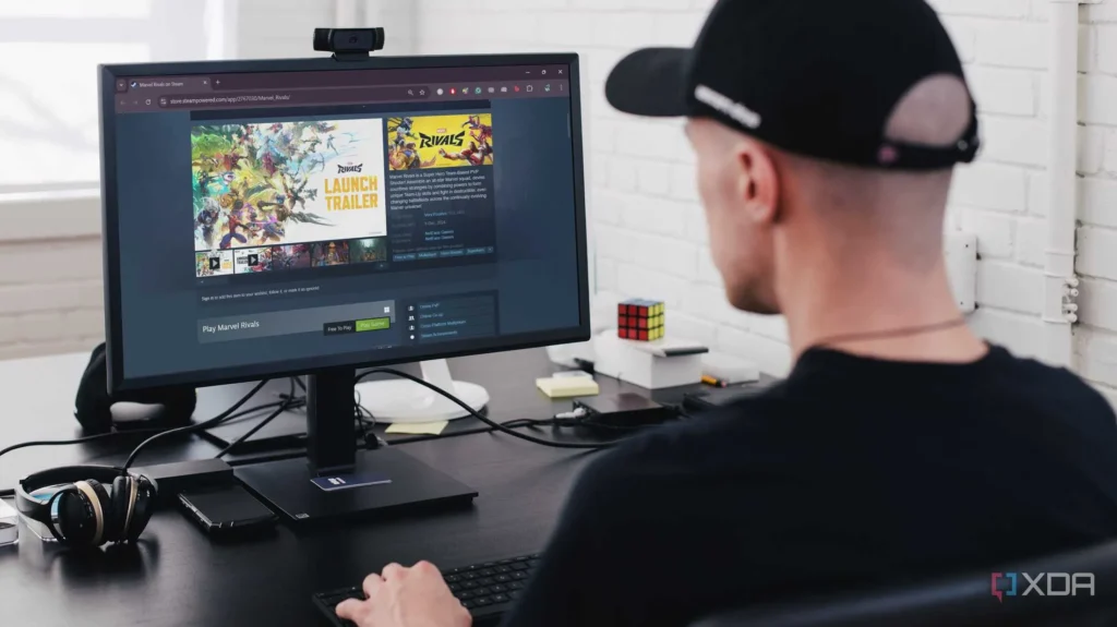 The image shows a person sitting at a desk in what appears to be a home office. In the foreground is a computer monitor displaying the Steam store page for a game called Marvel Rivals, and a keyboard. There is a pair of headphones to the left of the keyboard and a smartphone next to it. The background includes a partially visible white brick wall, a Rubik's Cube, and some other office supplies on the desk, suggesting a workspace setting.