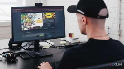 The image shows a person sitting at a desk in what appears to be a home office. In the foreground is a computer monitor displaying the Steam store page for a game called Marvel Rivals, and a keyboard. There is a pair of headphones to the left of the keyboard and a smartphone next to it. The background includes a partially visible white brick wall, a Rubik's Cube, and some other office supplies on the desk, suggesting a workspace setting.