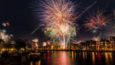 The image shows a vibrant fireworks display over a canal in Amsterdam, Netherlands. In the foreground, there are houseboats and the calm waters of the canal reflecting the colorful lights of the fireworks. The background is filled with the stunning spectacle of fireworks exploding in the night sky, with the city's buildings and trees lining the canal visible amidst the bursts of light. The city lights illuminate the buildings along the canal, creating a warm, inviting atmosphere against the dark night sky.