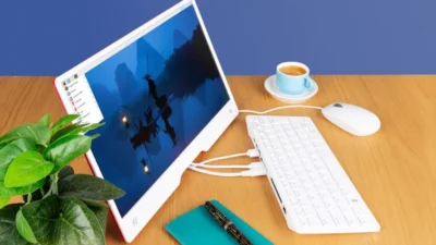 The image shows a workspace featuring a small, white computer monitor with a red back, a wireless keyboard and mouse, and a small potted plant. The monitor displays a scenic picture of a body of water with mountains in the background, perhaps a digital painting or a photograph. A cup of coffee sits on a saucer next to the monitor, adding to the relaxed atmosphere. All items are placed on a light brown wooden desk, suggesting a home office or personal workspace.