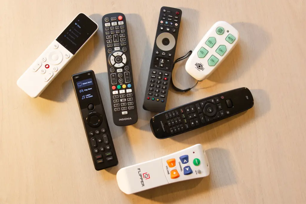 The image shows seven different TV remotes scattered on a light beige wooden table. In the foreground, there's a variety of remotes, each with unique designs and button layouts. The background is blurred but shows a consistent wooden surface. Several of the remotes display signs of use, with some buttons appearing more worn than others.