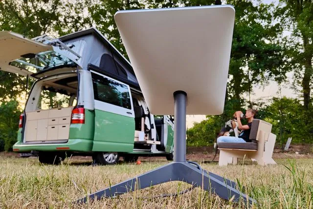 The photo shows a light green camper van parked in a grassy area, with lush green trees forming a backdrop. The van's rear doors are open, revealing a neatly organized interior. In the foreground, a portable satellite dish takes centre stage, its rectangular white dish is flat and pointing up at the sky. A small foldable chair is visible to the right, near a person sitting and petting a dog.