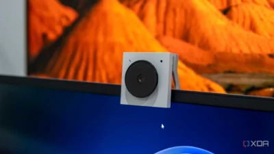 The image shows a close-up view of a small, square webcam attached to the top of a computer monitor. The webcam is white with a black circular lens. The monitor displays a blurry image in the background; it seems to be an abstract landscape of orange and brown hues, possibly a digital painting or a photograph of a desert-like environment. The foreground is dominated by the webcam and the top edge of the monitor, which is dark gray or black.