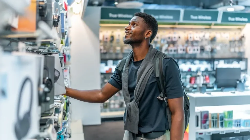 A man with short hair, wearing a dark blue t-shirt, a grey cardigan, and a backpack. He appears to be in his late twenties or early thirties. He is standing in an electronics store, with his right hand outstretched towards a box with headphones in, which are hanging on the wall.