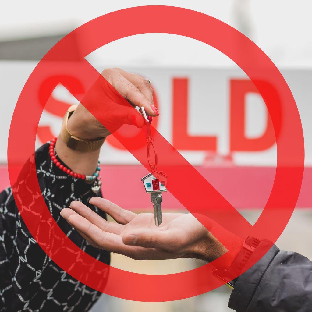 The image shows a house key being offered and being accepted by another. There is a large red circle and line superimposed on the image, symbolizing a prohibition or cancellation of such a transaction. In the foreground are two hands, one offering a key with a small house charm, and the other trying to receive it. The background features a blurry but clearly visible "SOLD" sign, suggesting a real estate transaction. The colour of the “SOLD” sign seems to be purposefully off, giving it a faded look, as if it's been there for a while.