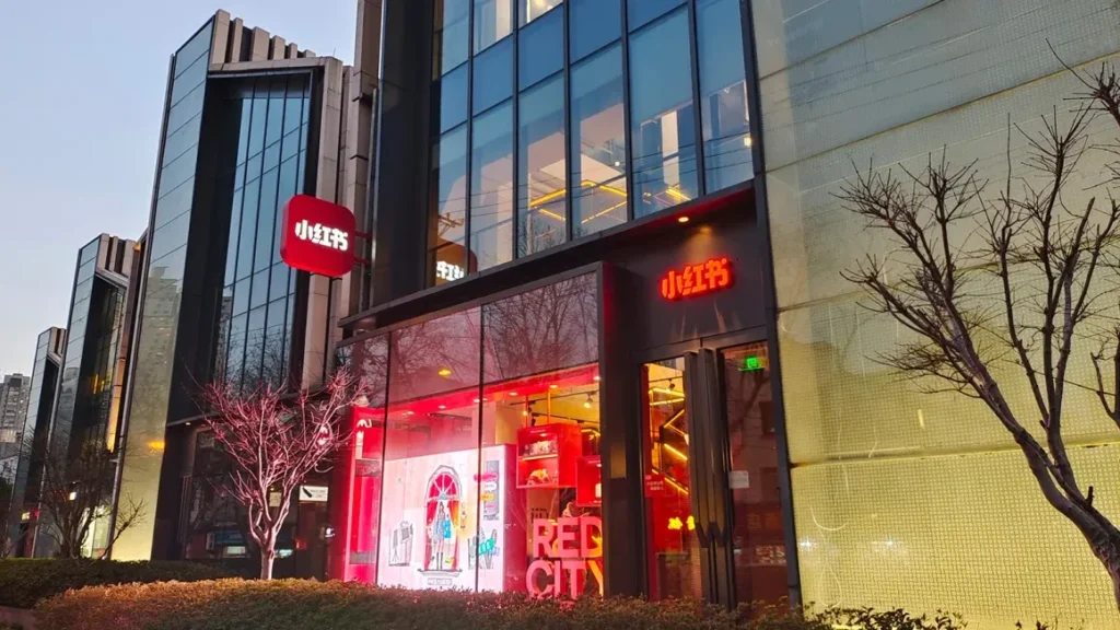 Modern storefront with large glass windows, red interior lighting, and signs featuring Chinese characters. Leafless trees and shrubs line the sidewalk.