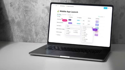 Laptop with screen open. On the screen is a project name saying Mobile App Launch. It has a list of tasks below it, and various priorities, progress, dates, and photos assigned to each task.