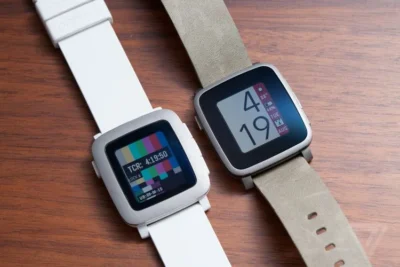 Two Pebble watches side by side on a wooden surface. One shows something that looks like a colour test pattern, and the other shows the time as 4 19 with the date and day of the week.