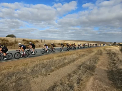 A group of cyclists racing down a wide, open road under a partly cloudy sky, with dry grass on either side.