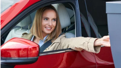 A woman in a beige coat extends her arm from the window of a red car, interacting with a drive-thru parking access service.