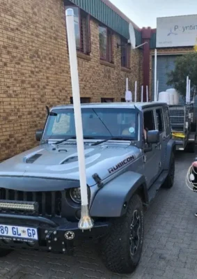 A gray Jeep Rubicon parked alongside a brick building, featuring tall antennas on its roof and a trailer behind it.