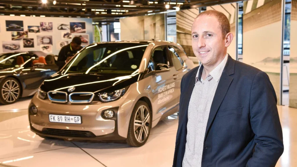 A stylish electric BMW i3 is showcased in a modern exhibition space, with sleek cars and promotional images in the background.